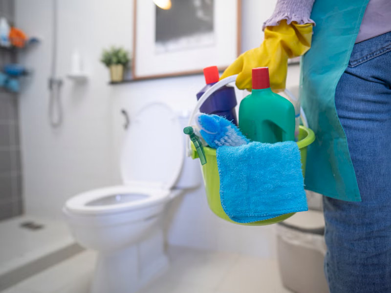 Người phụ nữ đeo găng tay vàng, mang xô đựng đồ dùng vệ sinh nhà tắm, chuẩn bị dọn dẹp bồn cầu trắng.