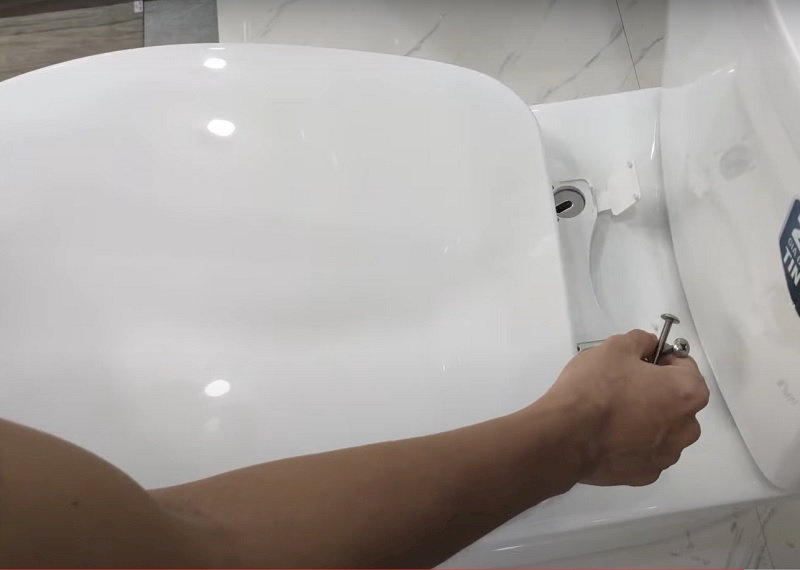 Người sửa bồn cầy đang dùng tay siết ốc vít bản lề nắp bồn cầu Inax trắng.