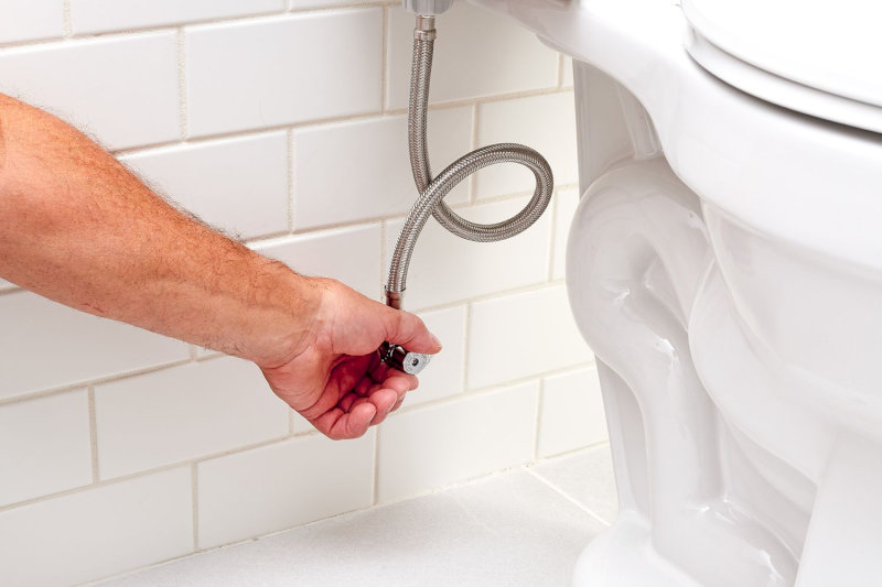 Người dùng đang siết tay nối dây cấp nước vào van dưới bồn cầu cạnh tường trắng