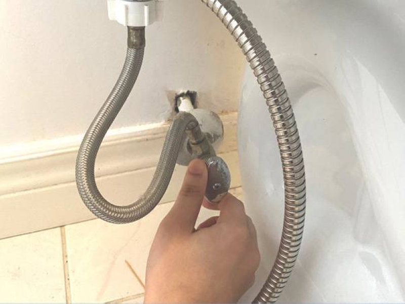  Tay người đàn ông đang vặn van kim loại để điều chỉnh hoặc khóa đường nước cấp cho vòi xịt bồn cầu.