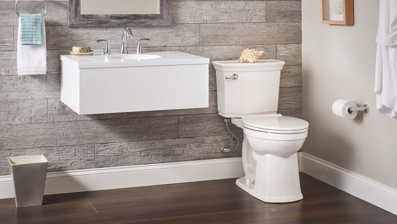Bồn cầu trắng đặt cạnh bồn rửa mặt treo tường màu trắng, tường ốp đá xám và sàn gỗ nâu.